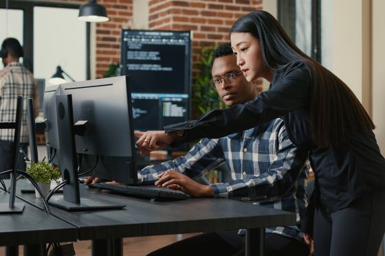 two people in an office reviewing code on a desktop setup with multiple monitors